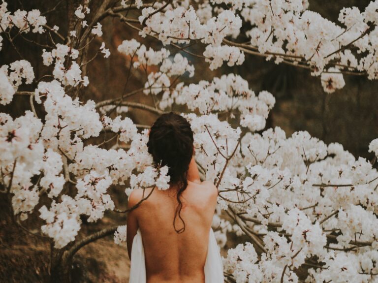 A woman with no clothes on facing a tree with white flowers.