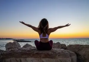 A woman sitting on a rock with both her arms out to each side during sunset.